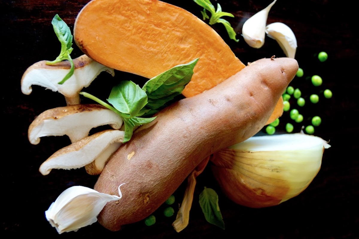 Half a sweet potato, fresh basil, garlic, onion, fresh basil, and peas on a black background.