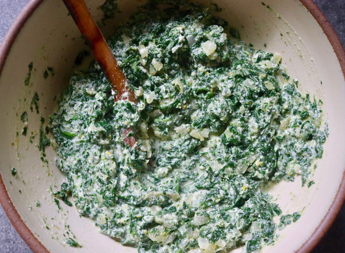 Cream-colored ceramic bowl with ingredients for filling for spinach and ricotta rolls all mixed together -- lemon zest and juice, Ricotta, spinach, Parmesan, onions and garlic and spices.