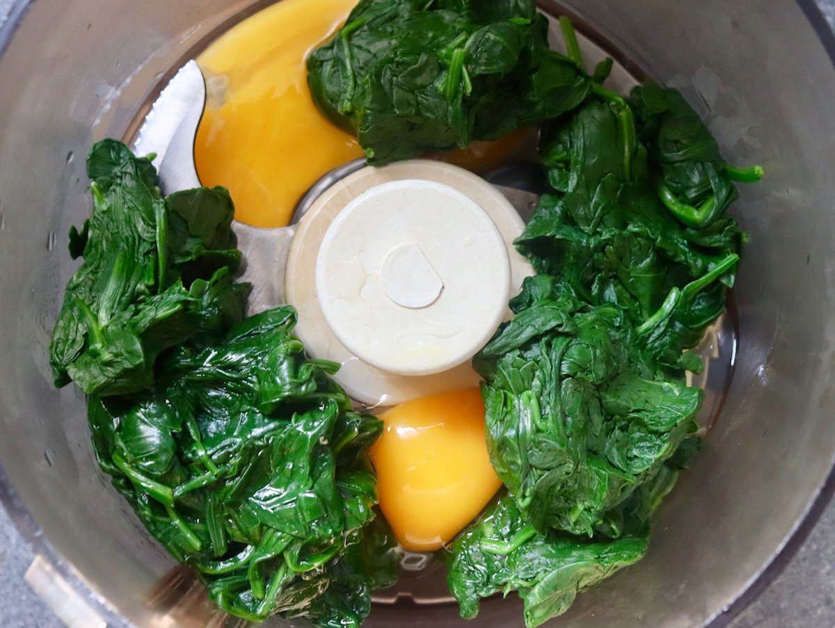 Cooked spinach and two eggs in a rood processor ready to be blended.