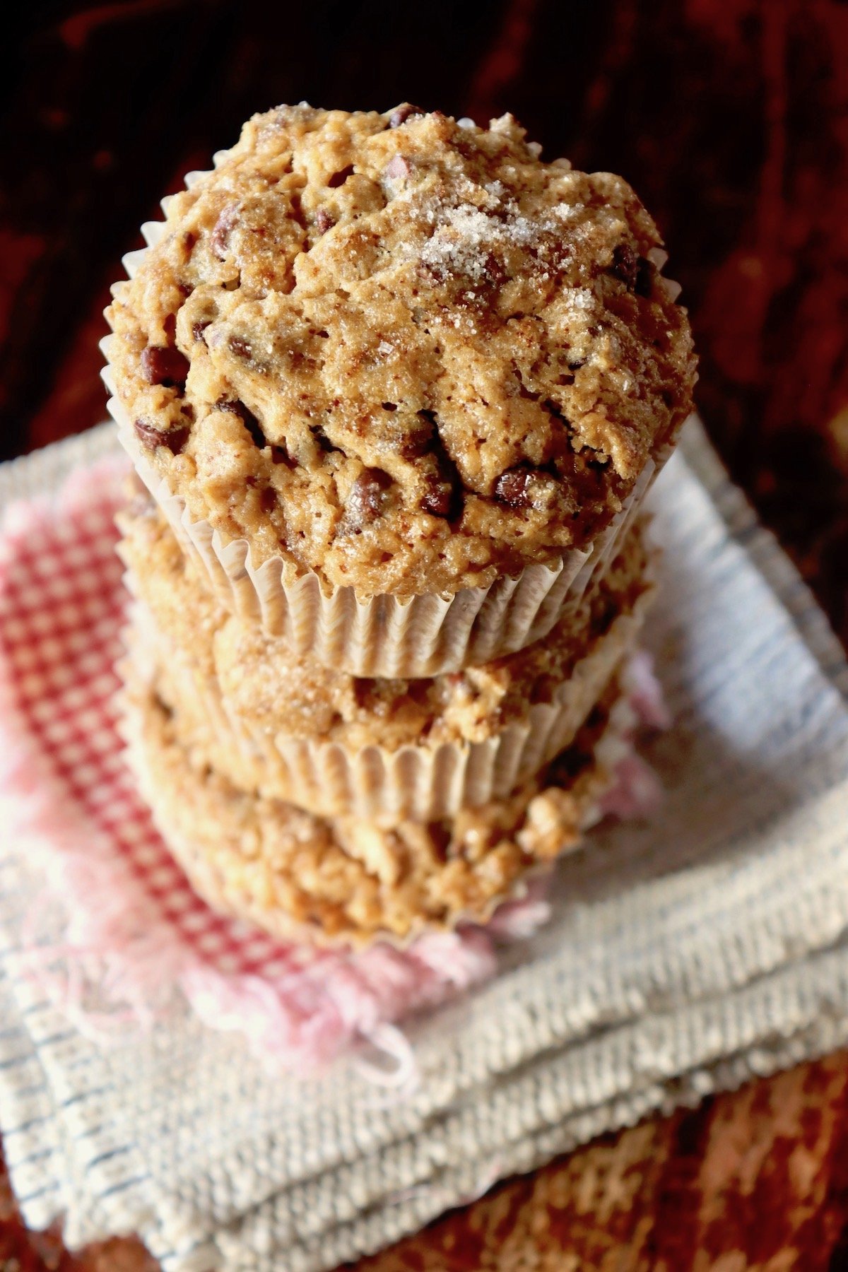 Stack of three cappuccino muffins in paper liners of stacks on beige and red cloth napkins.