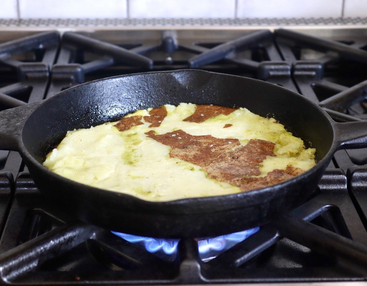 Large smashed Russet potato in cast iron skillet over high flame on stove.