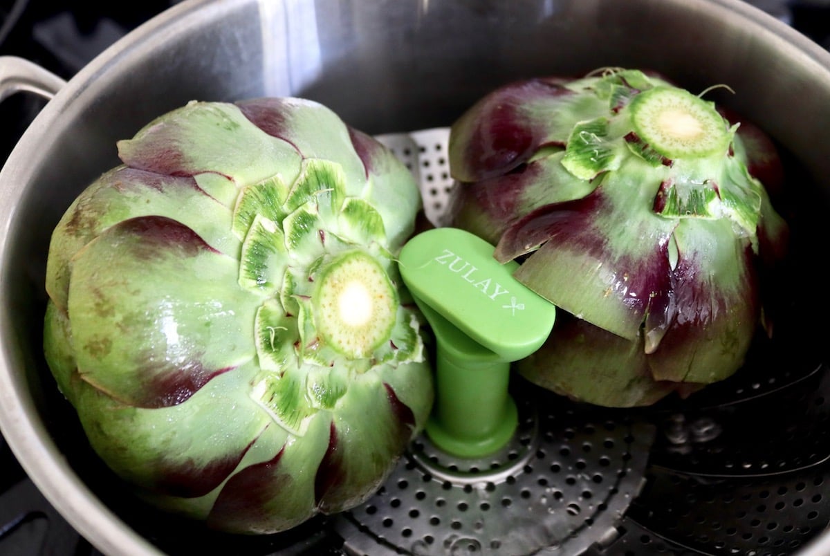 Two large green-purple, upside down artichokes in a steamer pot.