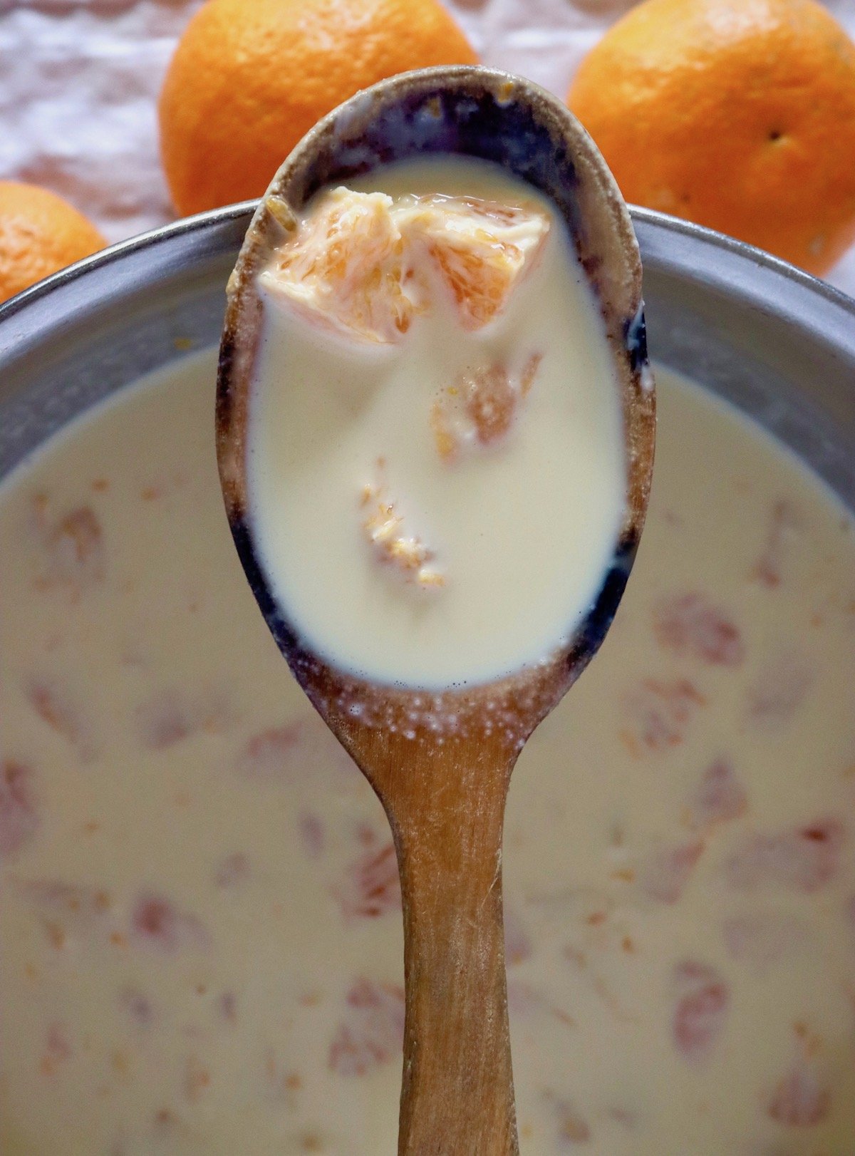 Wooden spoon with custard and tangerine pieces over a stainless steel bowl with more of it.