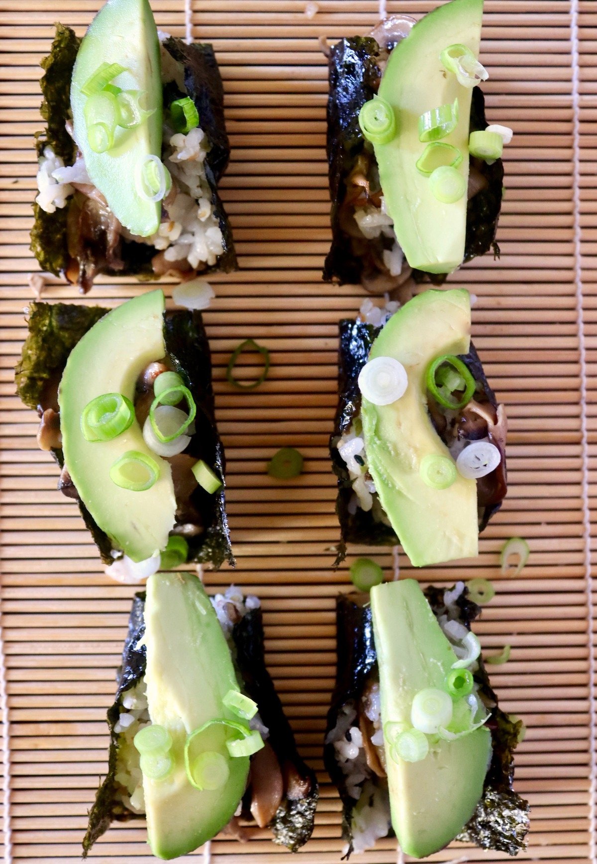 6 snack-sized Nori sheets on a sushi mat with rice-mushroom casserole and avocado inside.