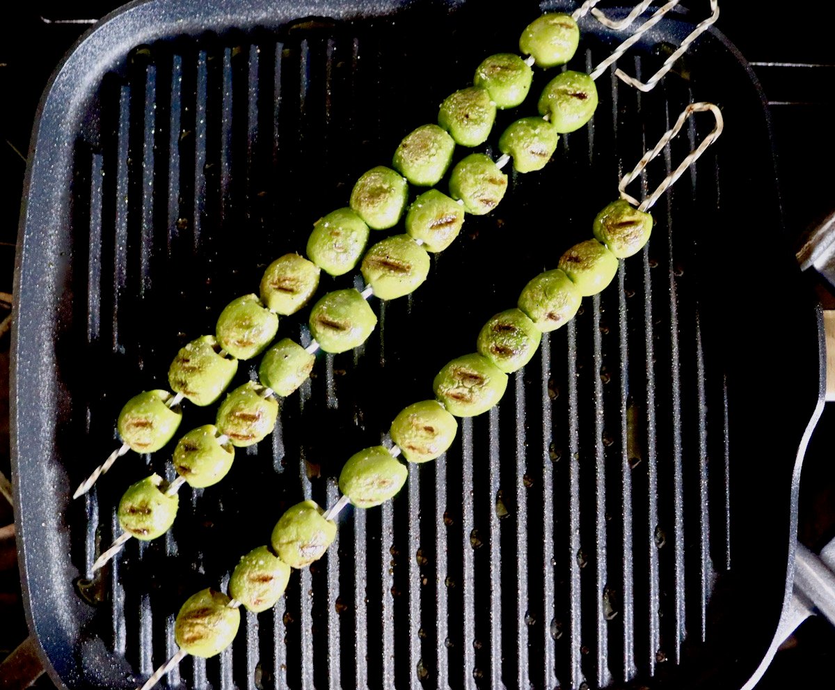 Three wire skewers full with grilled green olives on a black stove-top grill.