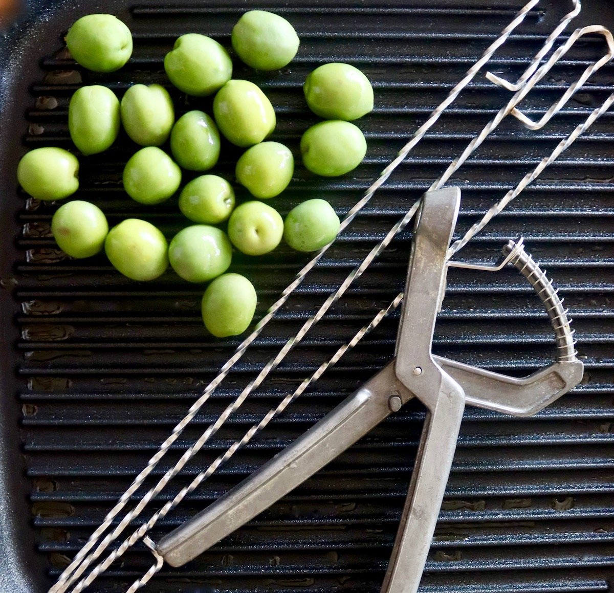 Black stove-top grill with olive pitter, green olives and three wire skewers.