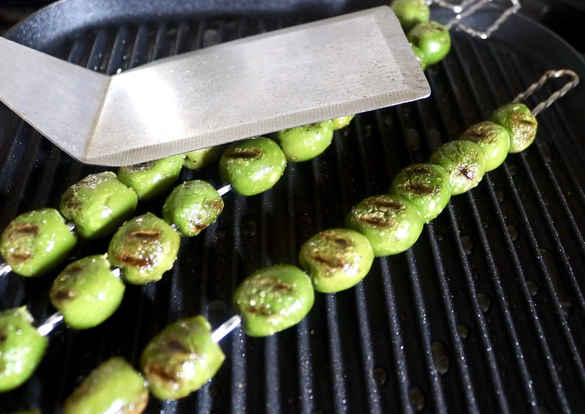 Large metal, flat-bottomed spatula gently pressing down on skewers with green grill olives, on black stovetop grill.