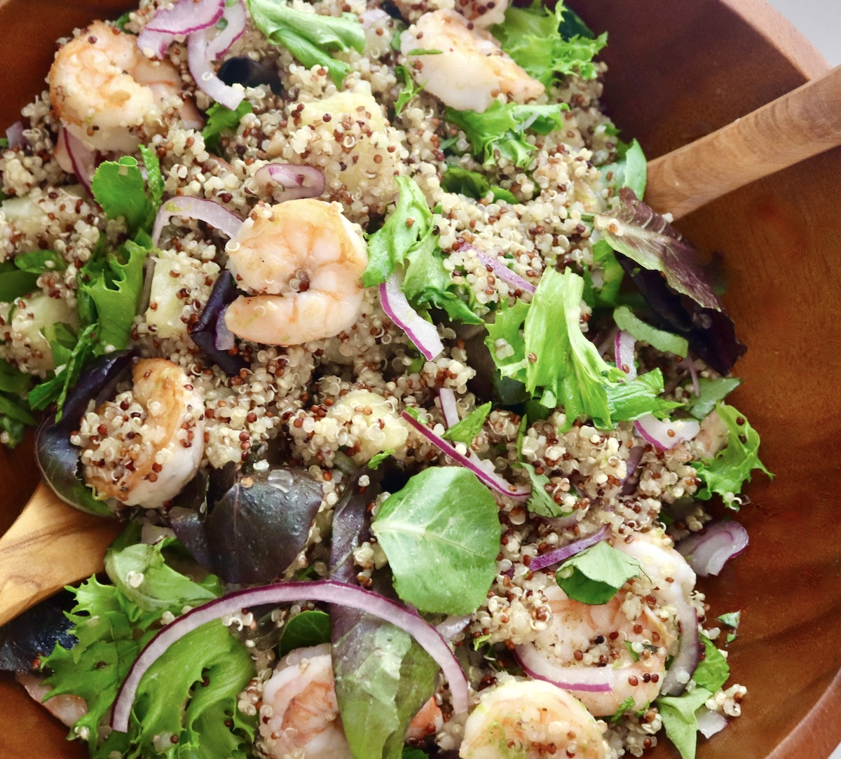 Large wooden salad bowl with mixed quinoa, cooked shrimp, sliced red onion, lettuce and fresh herbs.