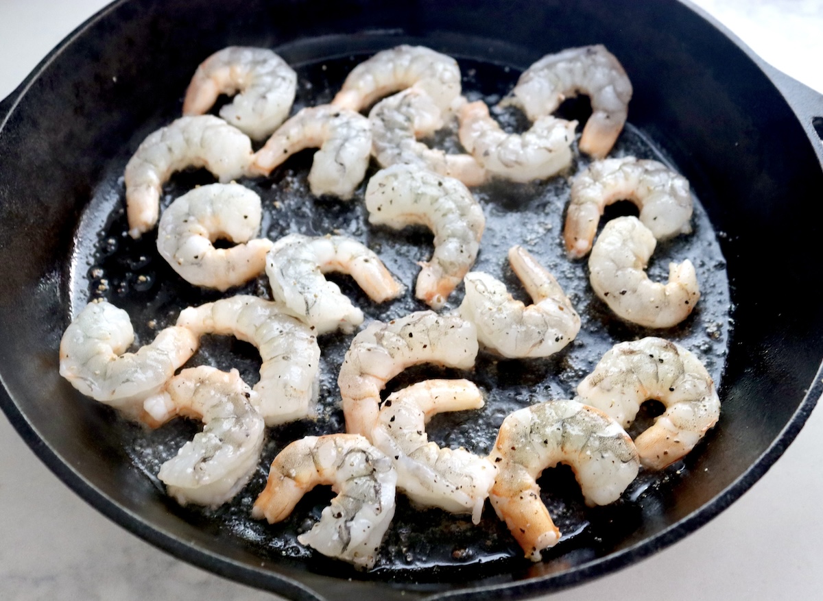 Several raw shrimp in a cast iron skillet, with slightly pink bottoms.