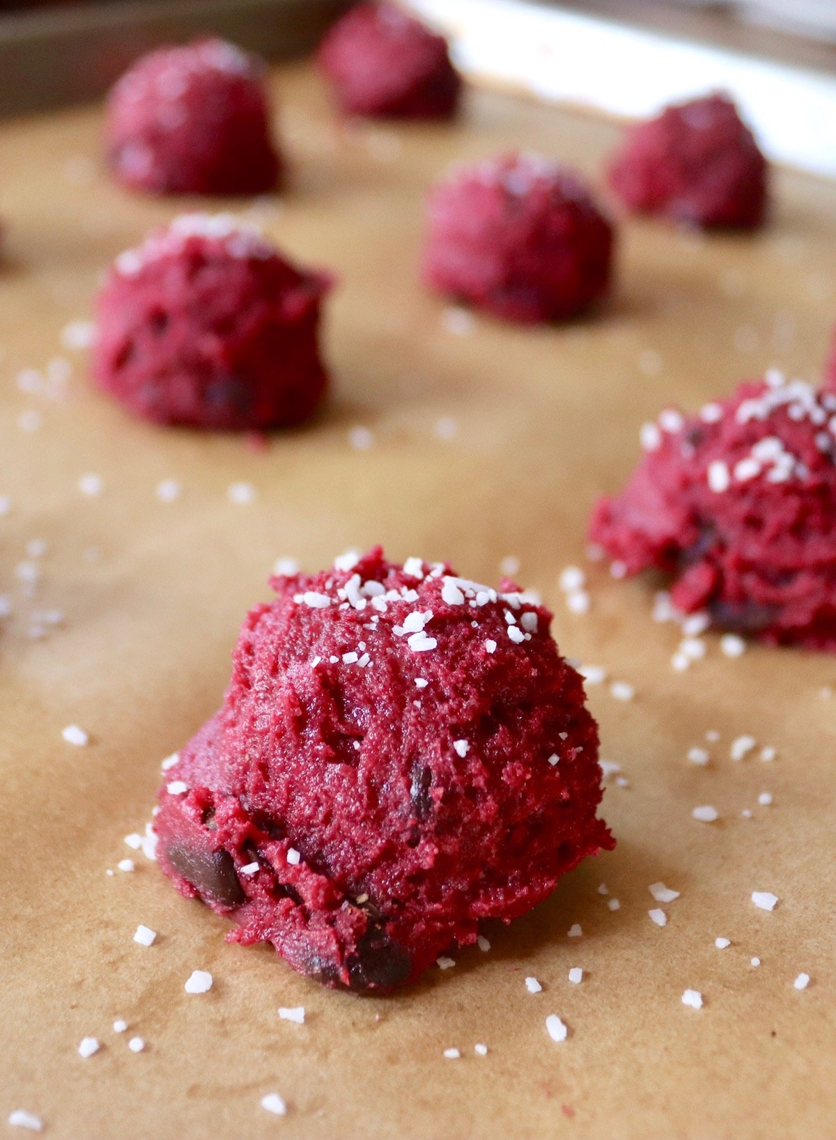 Chocolate chip beet cookie dough shaped into balls with sprinkled salt, on parchment paper.