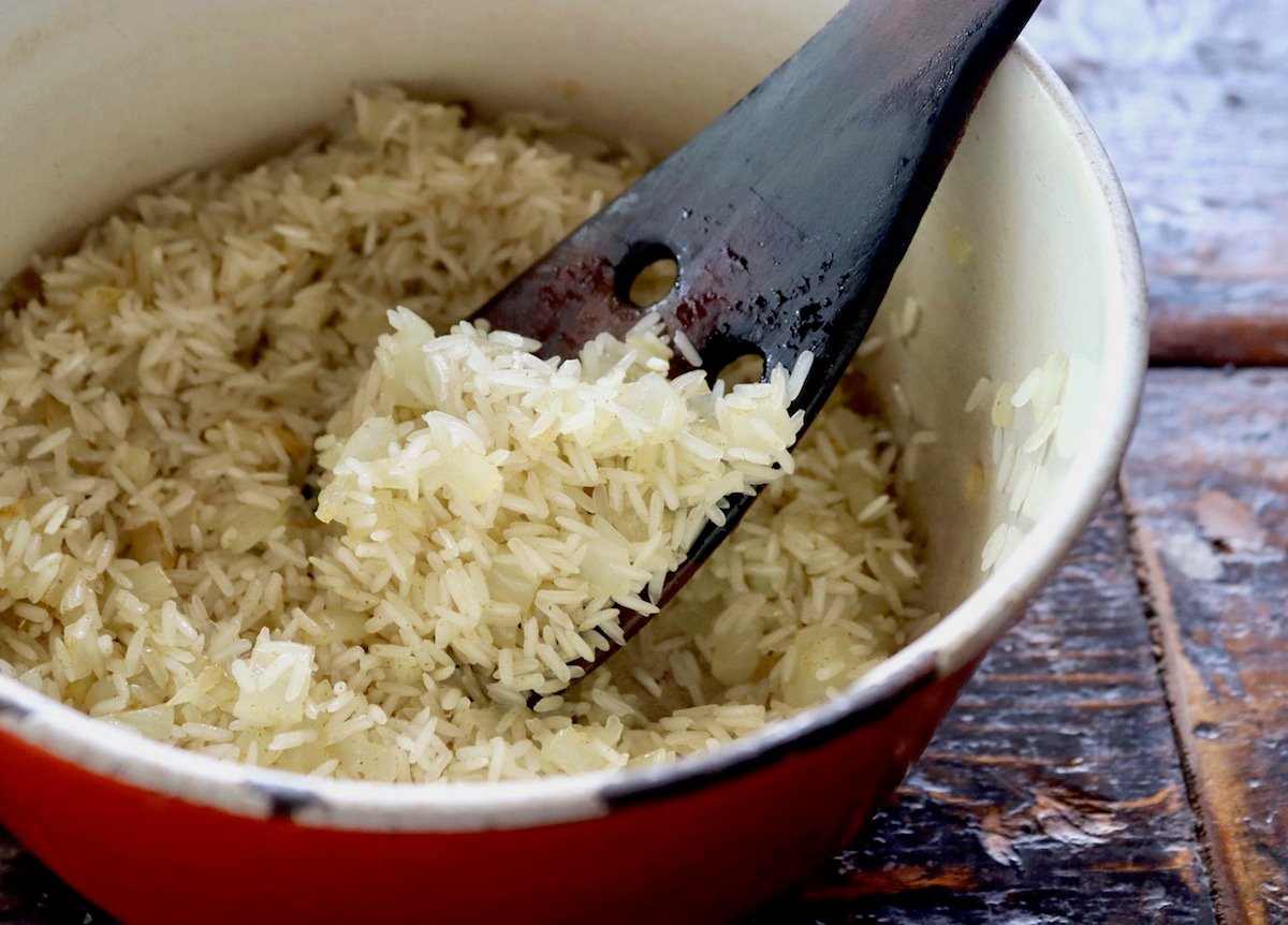 Rice mixed with chopped onion and oil in a pot with a wooden spatula.