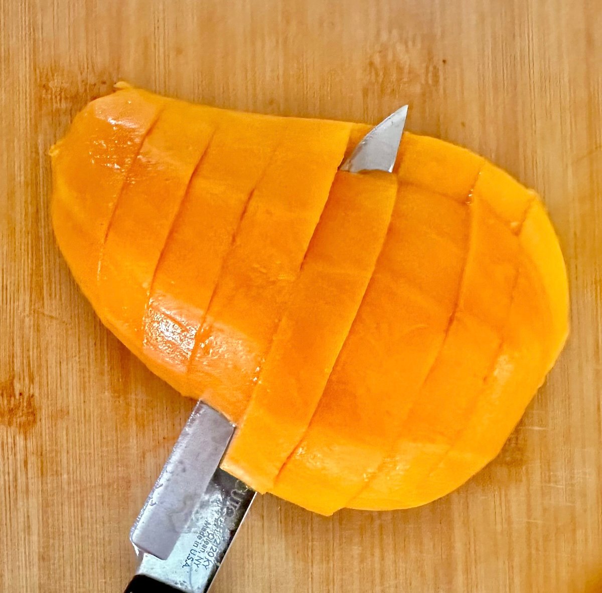 Peeled mango half being cut off the pit, on a light wood cutting board.