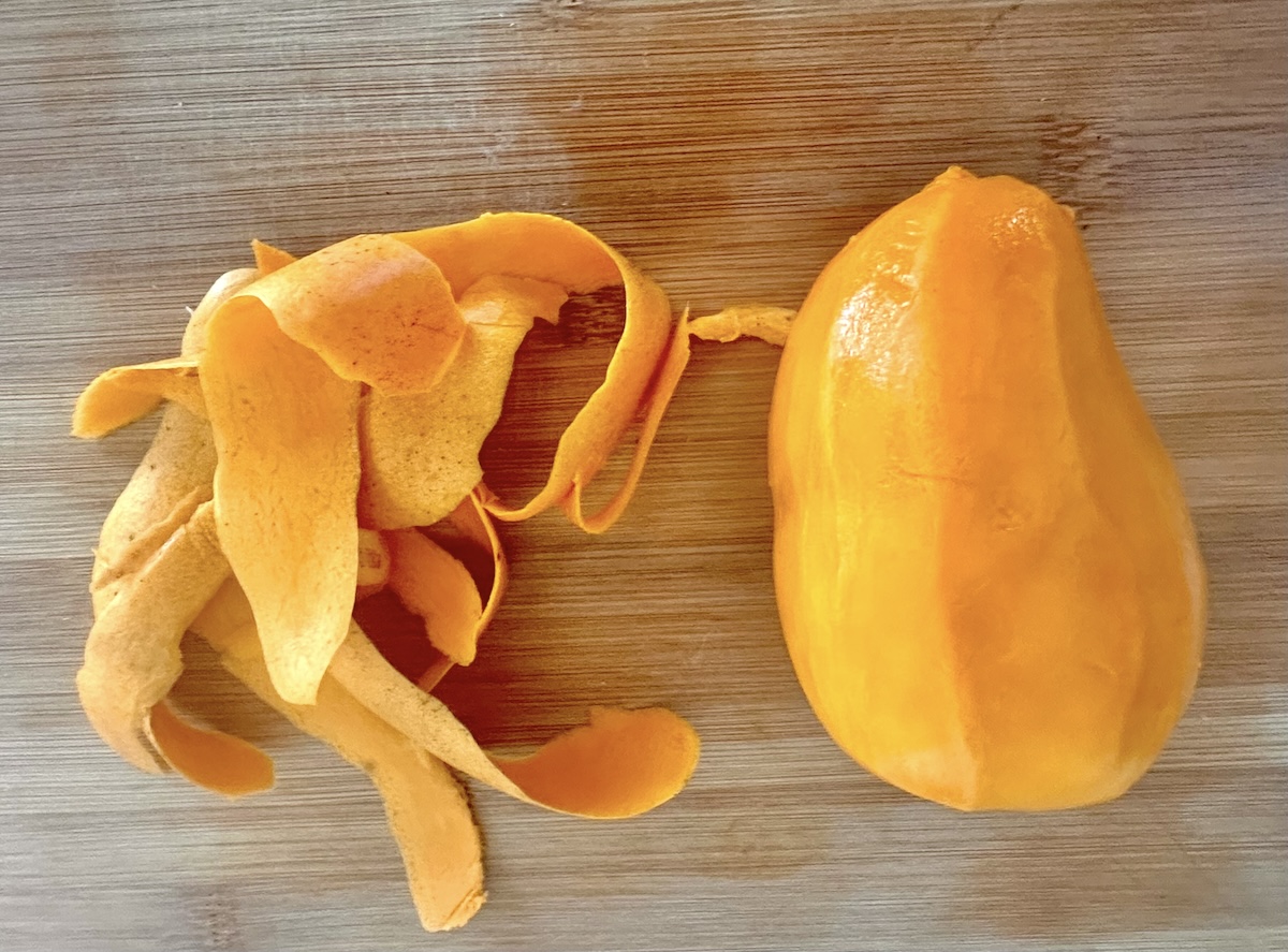 Peeled mango with its skin next to it on a light wood cutting board.