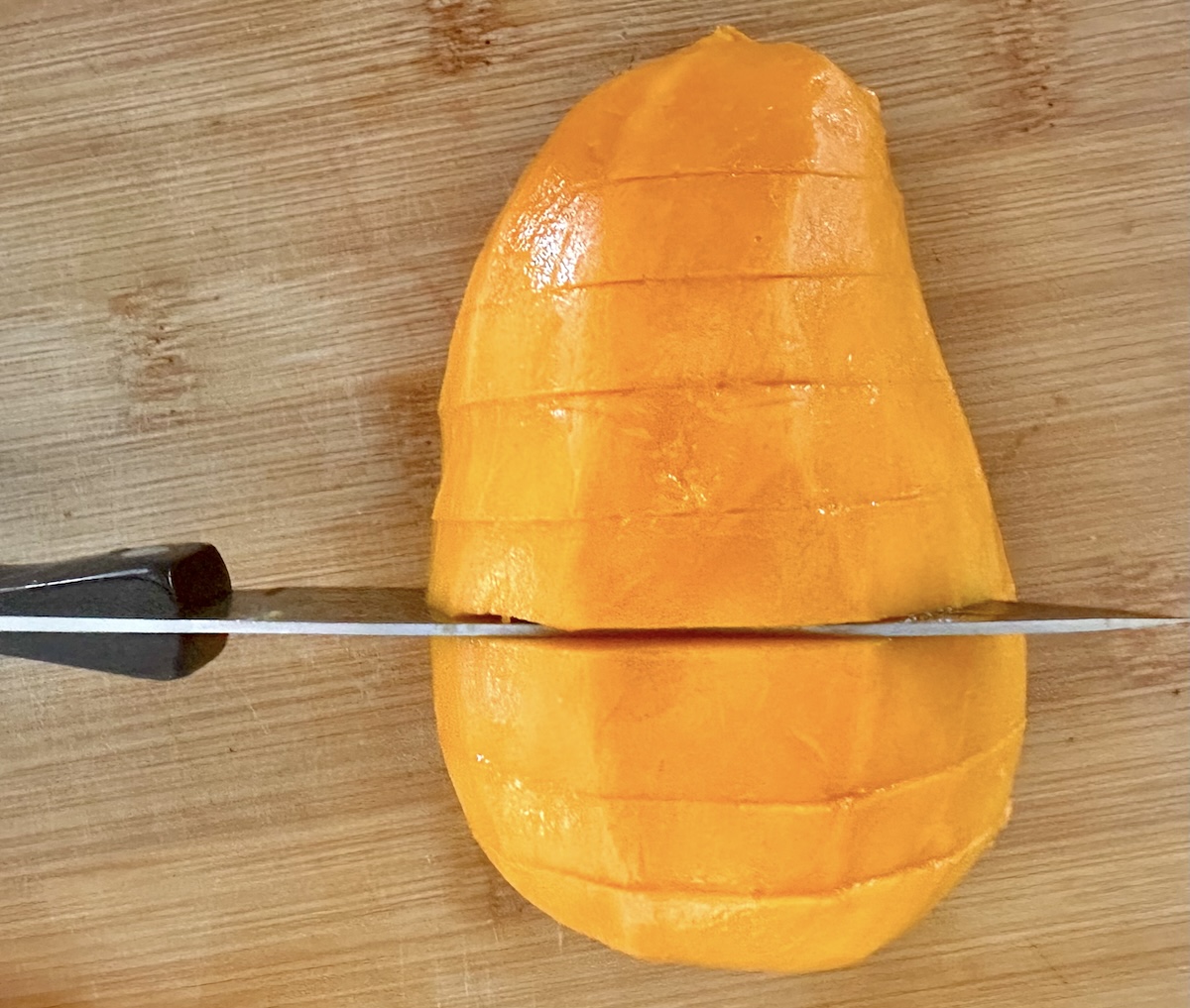 Peeled mango with a paring knife cutting horizontal slits across it, on a light wood cutting board.