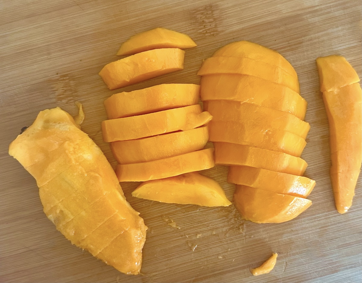Mango pit and peeled mango halves sliced into many pieces, on a light wood cutting board.