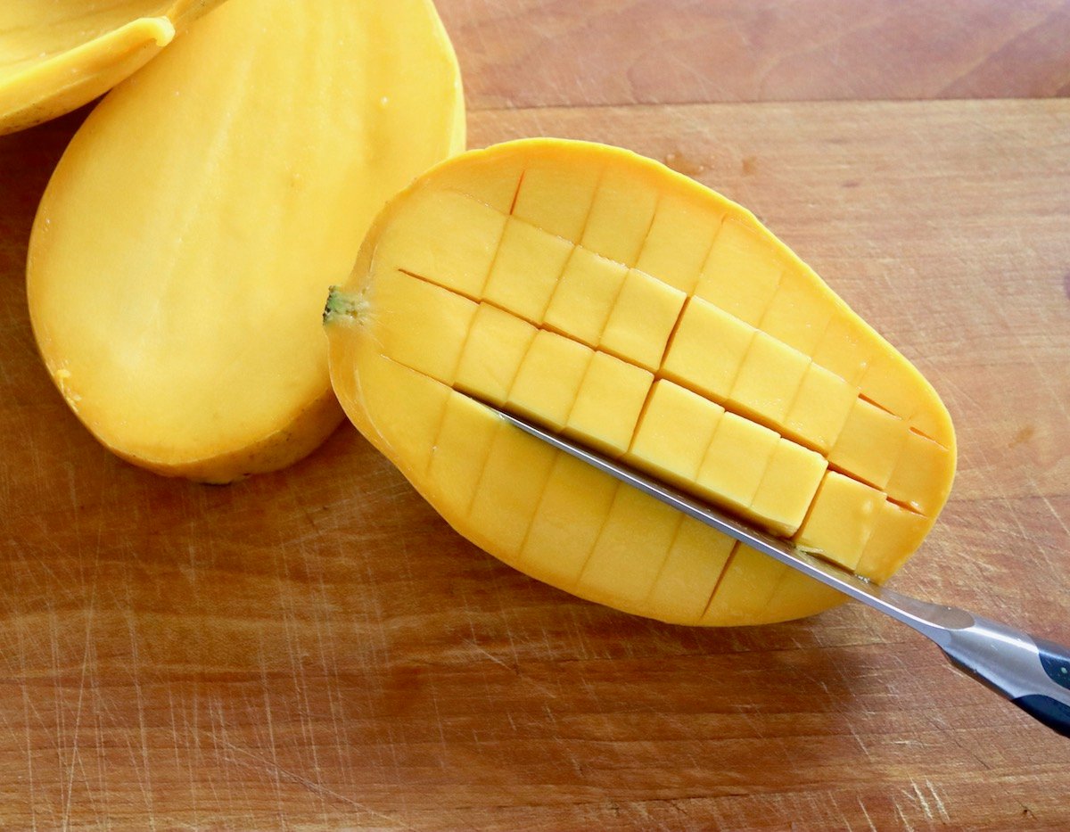 Mango sliced in half and diced within the skin with paring knife, on wooden cutting board.