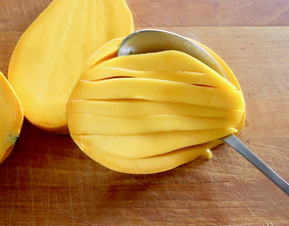 Mango sliced in half and sliced within the skin with a spoon scooping the pieces out, on wooden cutting board.