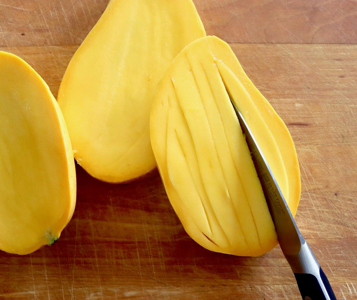 Mango sliced in half and sliced within the skin with paring knife, on wooden cutting board.