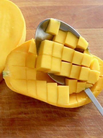 Mango sliced in half and diced within the skin with a spoon scooping the pieces out, on wooden cutting board.