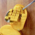 Mango sliced in half and diced within the skin with a spoon scooping the pieces out, on wooden cutting board.