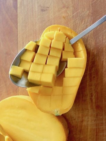 Mango sliced in half and diced within the skin with a spoon scooping the pieces out, on wooden cutting board.