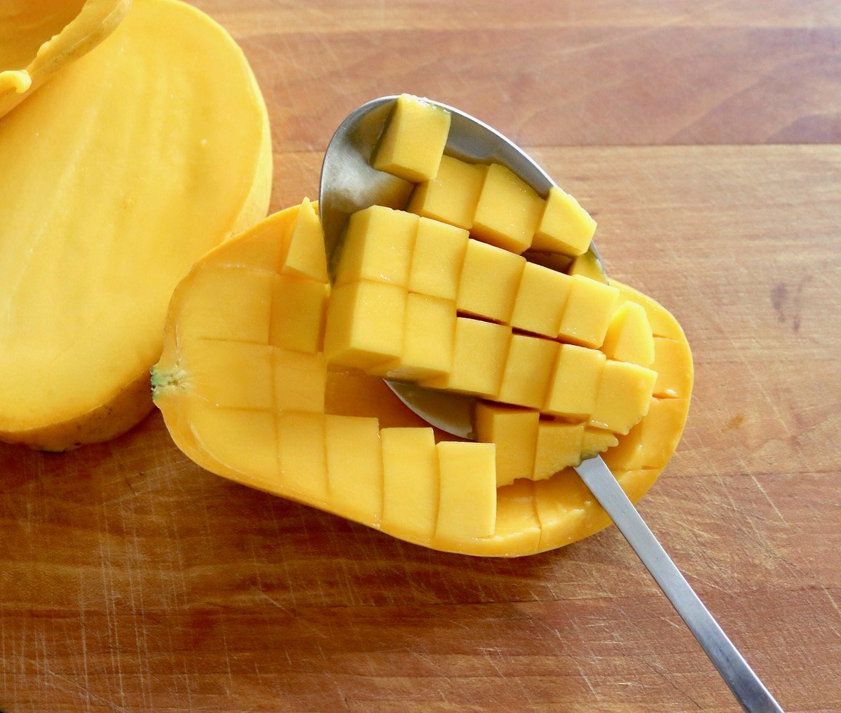 Mango sliced in half and diced within the skin with a spoon scooping the pieces out, on wooden cutting board.