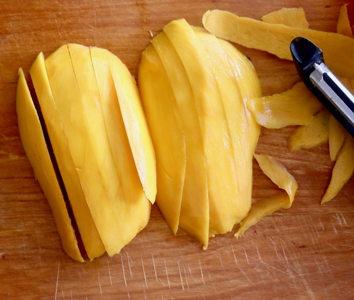 Two peeled mango halves cut into thin slices on cutting board.