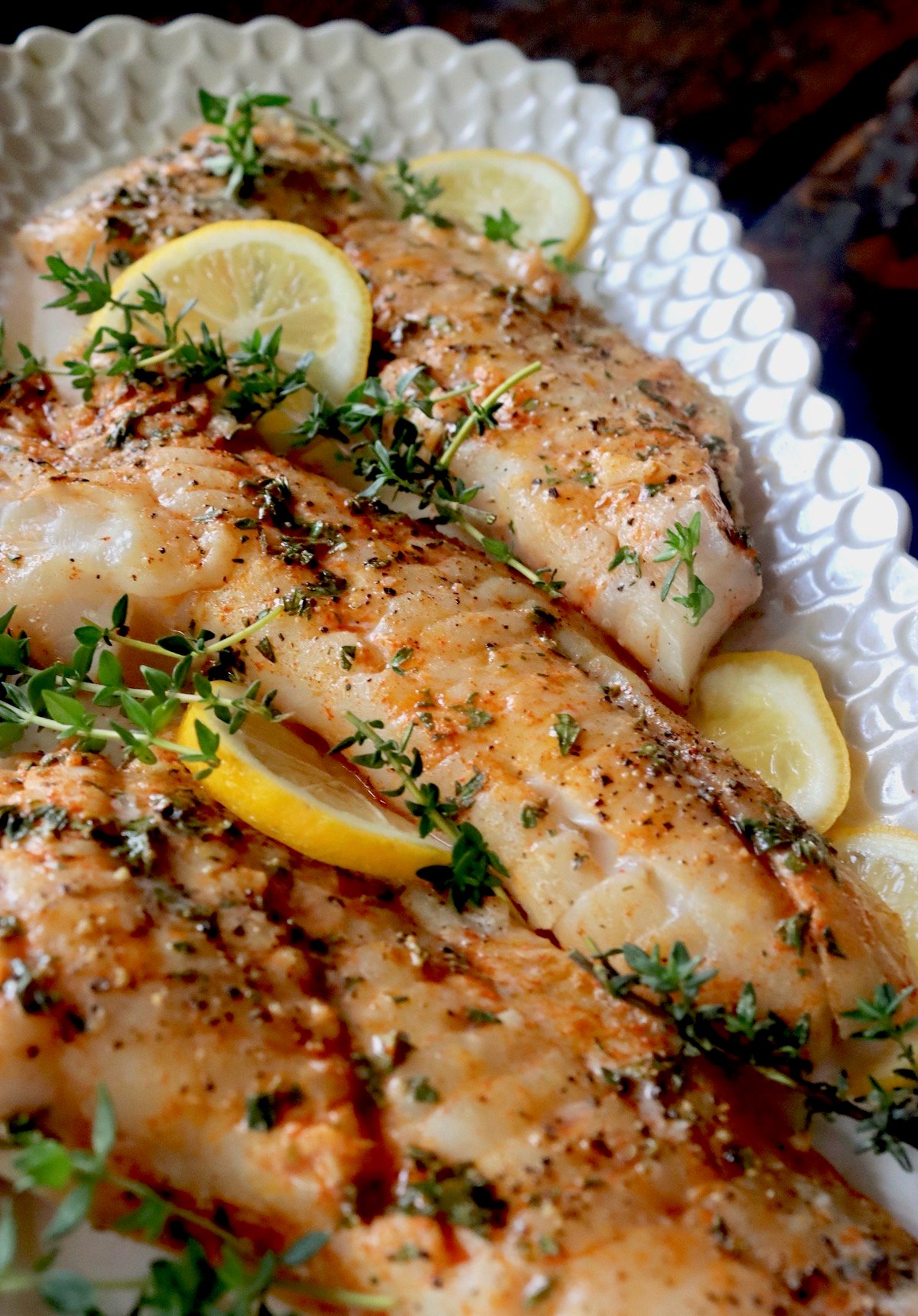 Three baked Rockfish Fillets on white platter with fresh thyme and lemon slices.