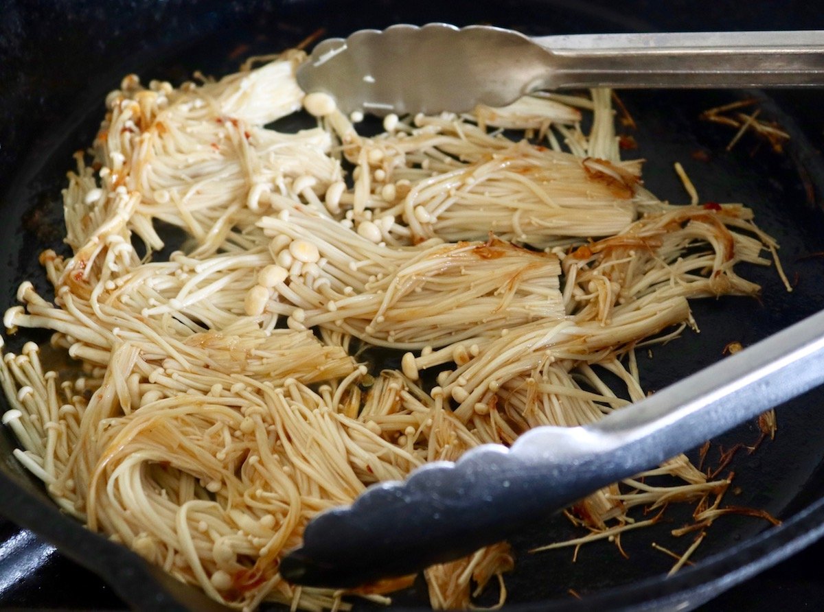 Enoki mushrooms slightly sauteed with metal tongs in a cast iron skillet.