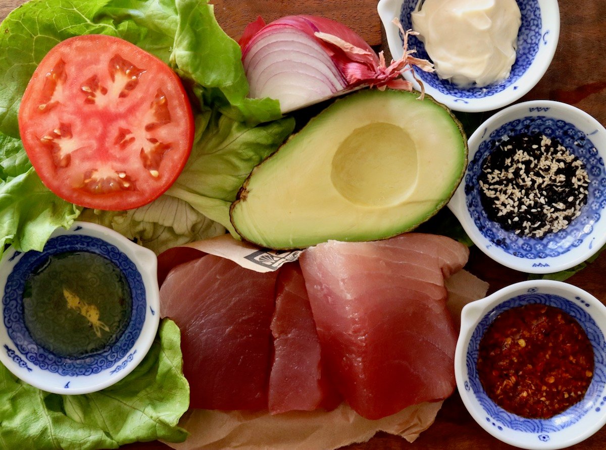 Ingredients for an ahi tuna burger including ahi tuna fillets, sesame seeds, avocado, red onion, tomato, lettuce and chili crunch.