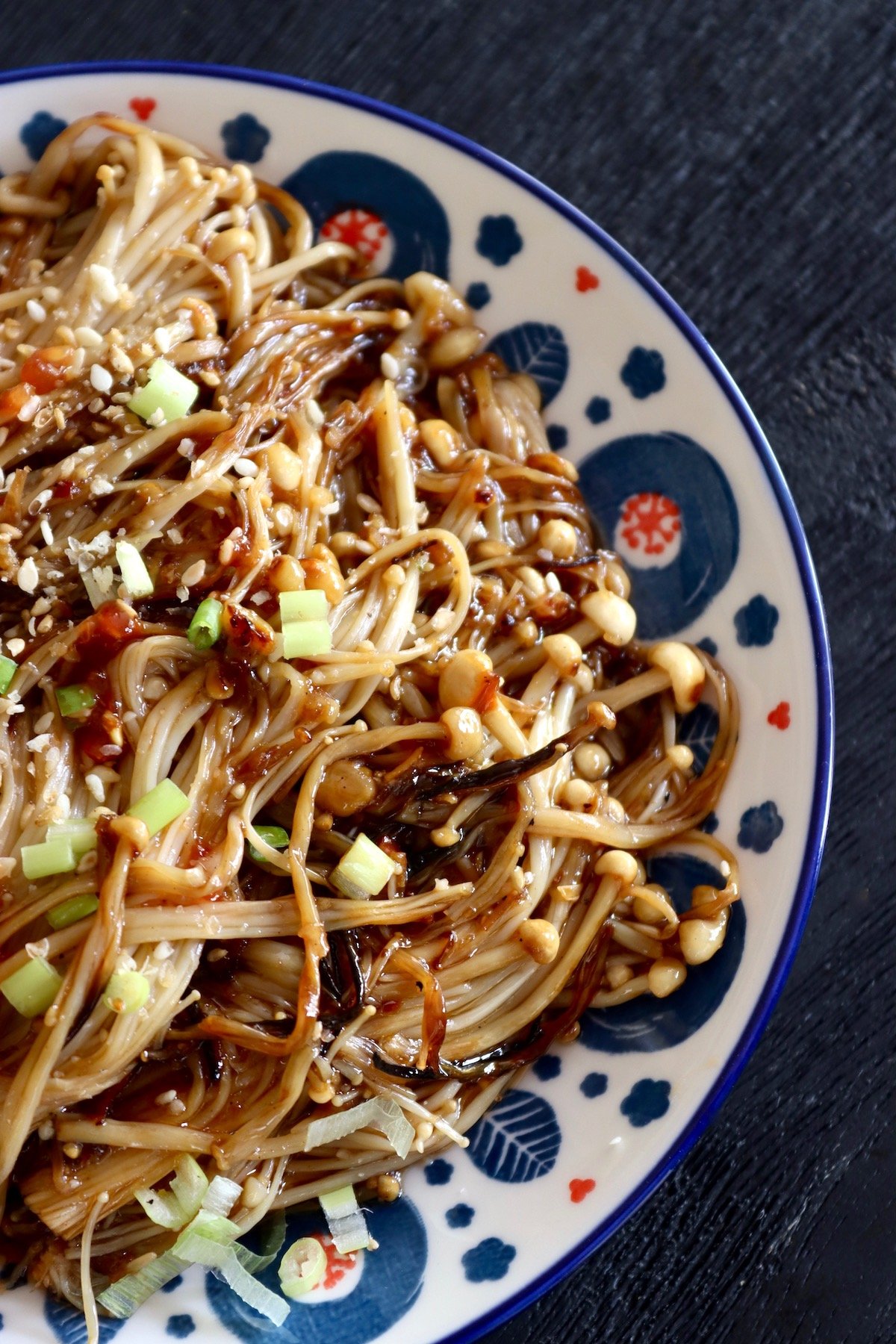 White plate with a blue and red dot-like pattern along the edges, full with enoki mushrooms in a spicy Asian sauce.