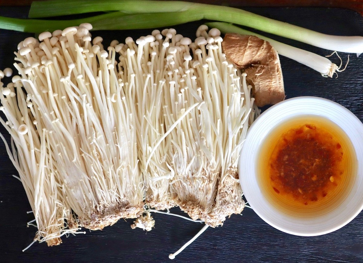 Three bunches of white Enoki mushrooms on a black tray with a small piece of ginger root, green onions and a tiny white bowl with red chili sauce.