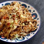 Close up of a white plate with a blue and red dot-like pattern along the edges, full with enoki mushrooms in a spicy Asian sauce.