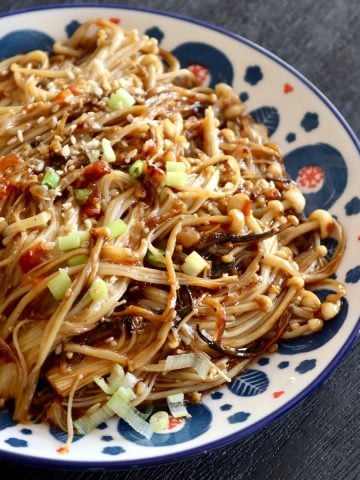 Close up of a white plate with a blue and red dot-like pattern along the edges, full with enoki mushrooms in a spicy Asian sauce.
