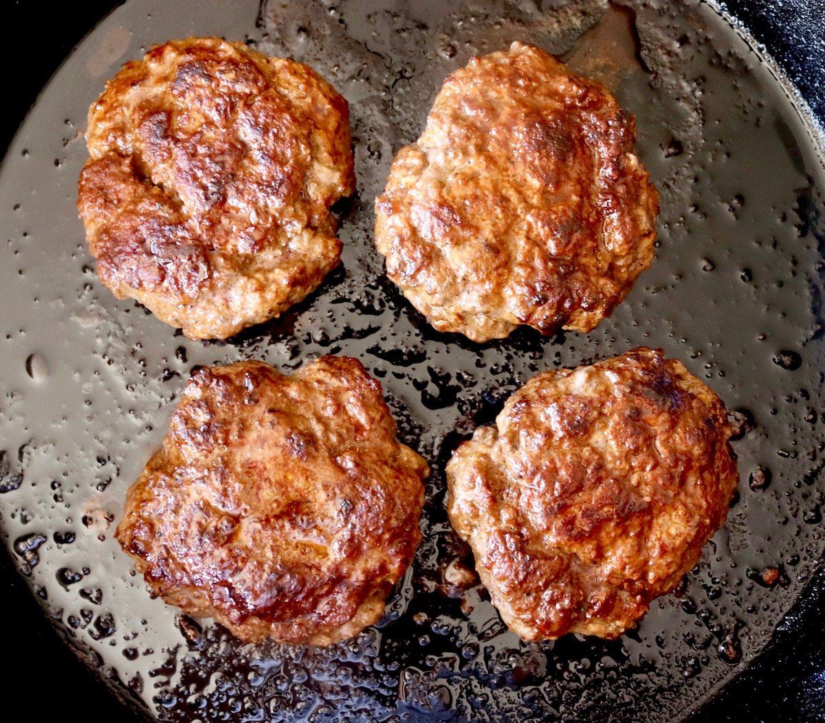 4 browned, cooked meat patties in a cast iron skillet.