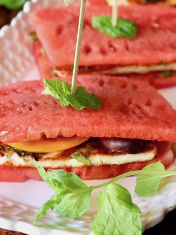 Close up of 2 watemelon Halloumi sandwiches on a white narrow plate with toothpicks and mint leaves.