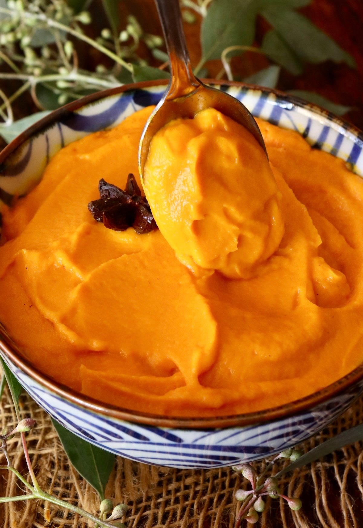 Carrot Puree in a blue and white bowl with on star anise pod on top and a large spoon scooping some out of it.