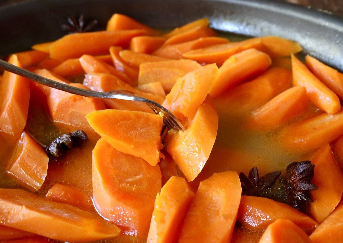 Cooked carrot slices with in buttery water and several Star Anise, in a sauté pan with a fork.