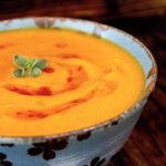 Bright orange Red Kuri Squash pureed soup, in a blue bowl with a small oregano leaf on top.