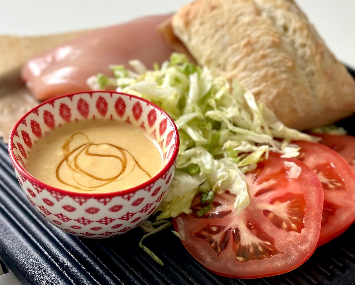 Small red and white bowl with honey mustard, shredded lettuce, bread, tomato slices and raw chicken sitting on a grill.