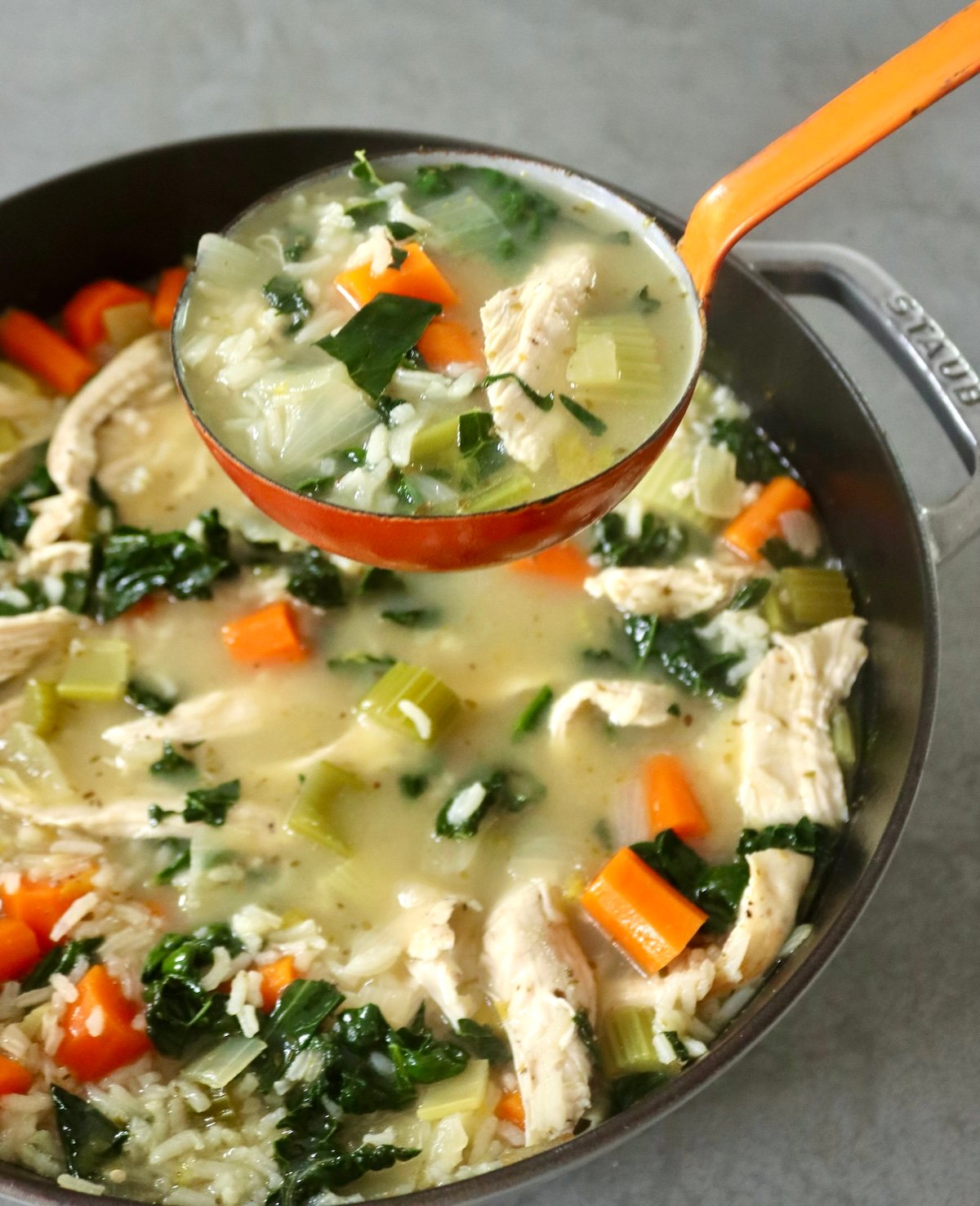 Chicken soup with vegetables held in an orange ladle with the soup pot below it.