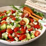 Salad with tomatoes and cucumber with wooden salad servers in white bowl.