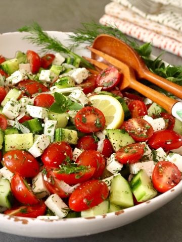 Salad with tomatoes and cucumber with wooden salad servers in white bowl.