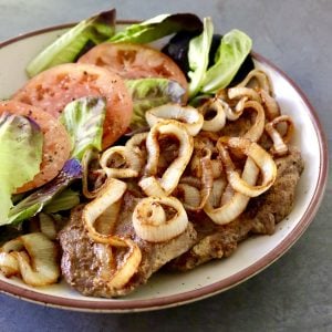 Round serving platter with steak and caramelized onion and tomato and lettuce next to it.