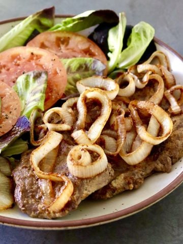 Round serving platter with steak and caramelized onion and tomato and lettuce next to it.