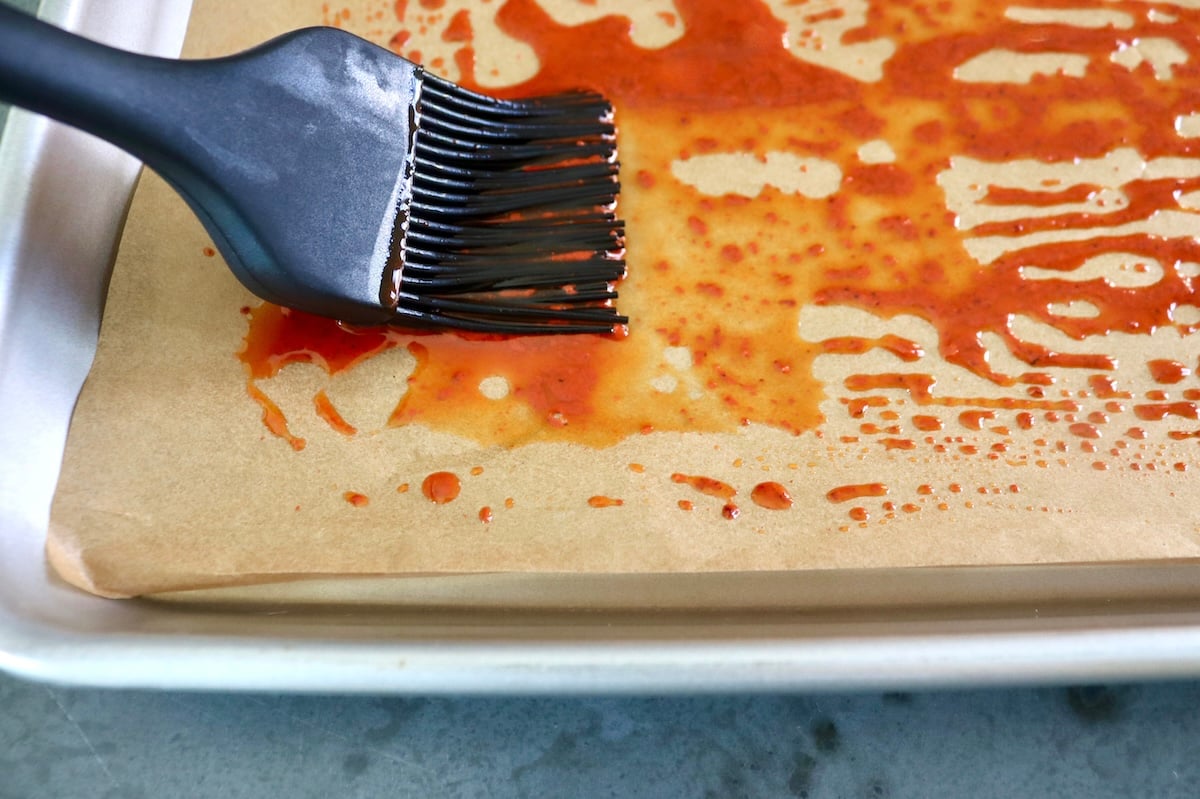 Achiote sauce being spread on a parchment-lined sheet pan with a black silicone brush.