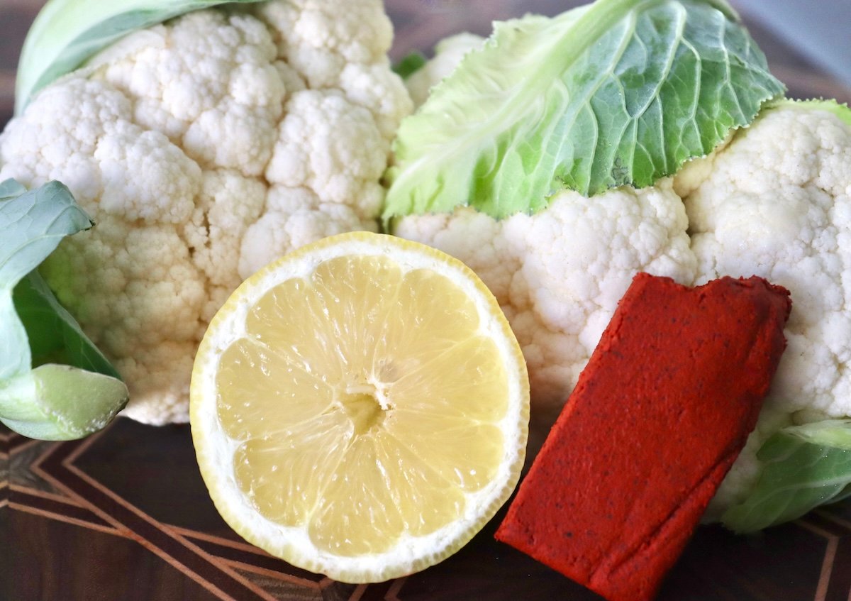 two small heads of cauliflower, achiote paste and half a lemon on a cutting board.
