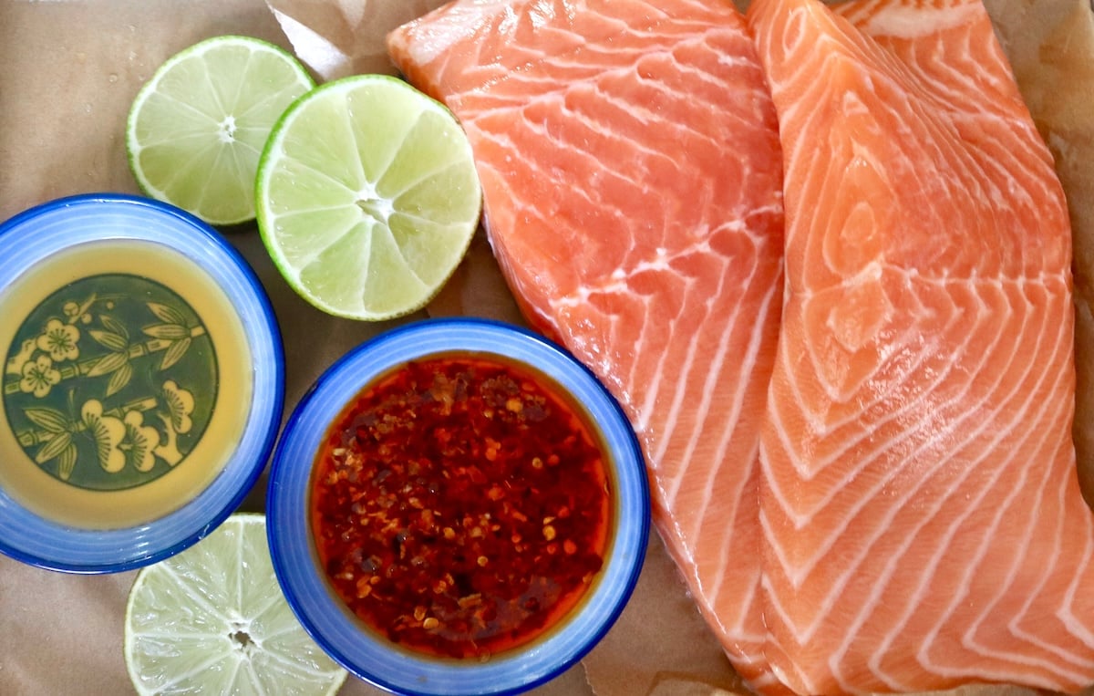 Two raw fillets of salmon with slices of lime and two tiny blue bowls - one with chili crunch and one with honey.