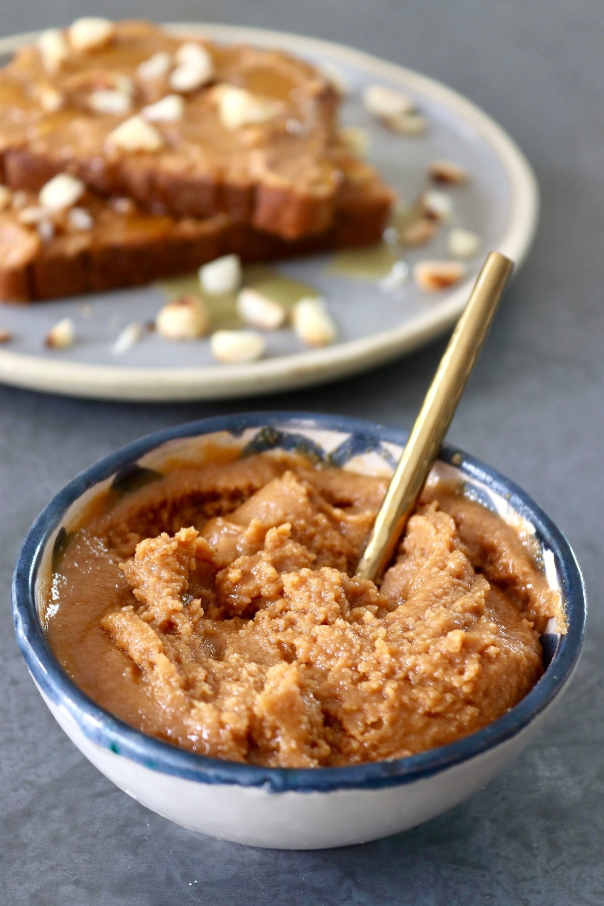 Macadamia-Nut-Butter Small dark-blue-rimmed bowl with macadamia nut butter and a place behind it with the butter spread on toast, with a few sprinkled whole nuts.