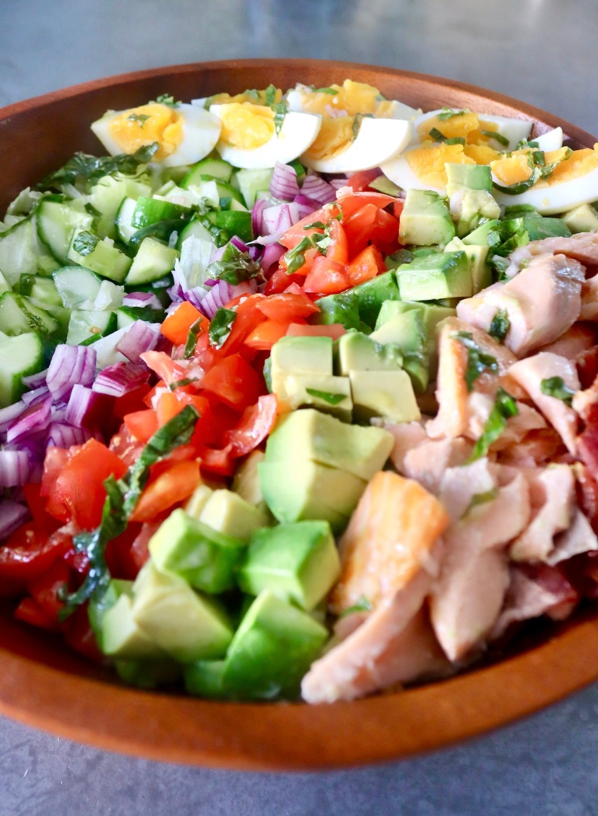 Wooden bowl filled with a composed salmon cobb salad, with rows of tomato, salmon, cucumber, red onion, basil, salmon and hard boiled eggs.
